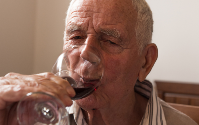 An elderly man with short gray hair is sitting indoors, drinking red wine from a glass. His face is wrinkled, and he is wearing a striped shirt under a sweater. The background is softly blurred, focusing on the man enjoying his drink.